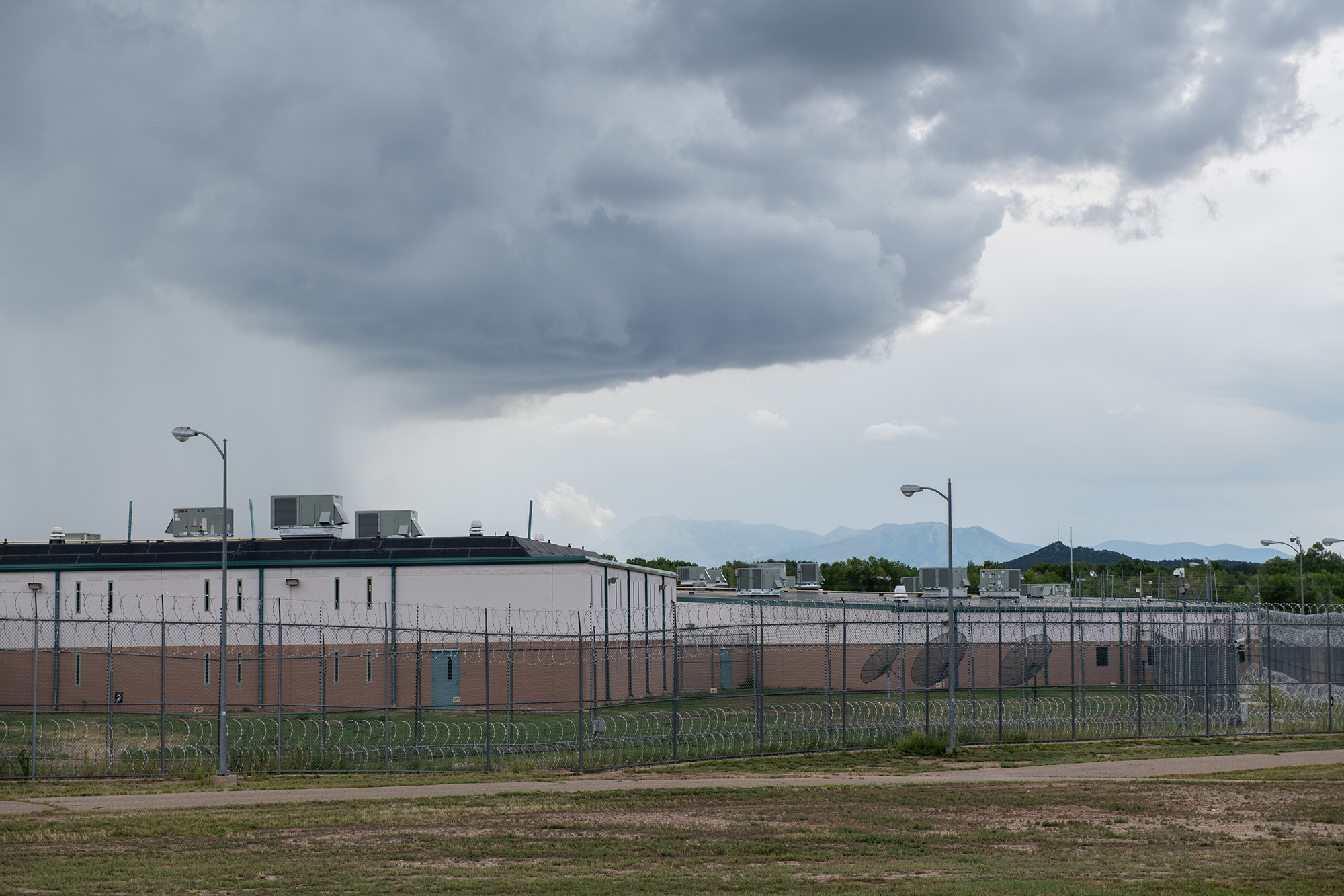 A squat prison surrounded by a barbed-wire fence, with a large, dark cloud hovering above it.