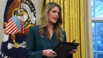 A woman with blond hair and heavy makeup, wearing a dark green suit, stands in the Oval Office holding a binder and pen.