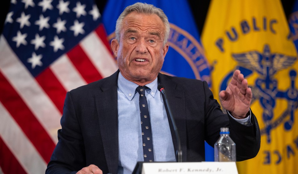 RFK Jr. with his hand raised, in front of flags, speaking