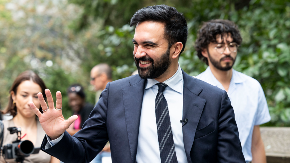 A young man with dark hair and a dark beard is dressed in a dark suit and dark tie with gray stripes. Outdoors in front of media, the man waves with a smile.
