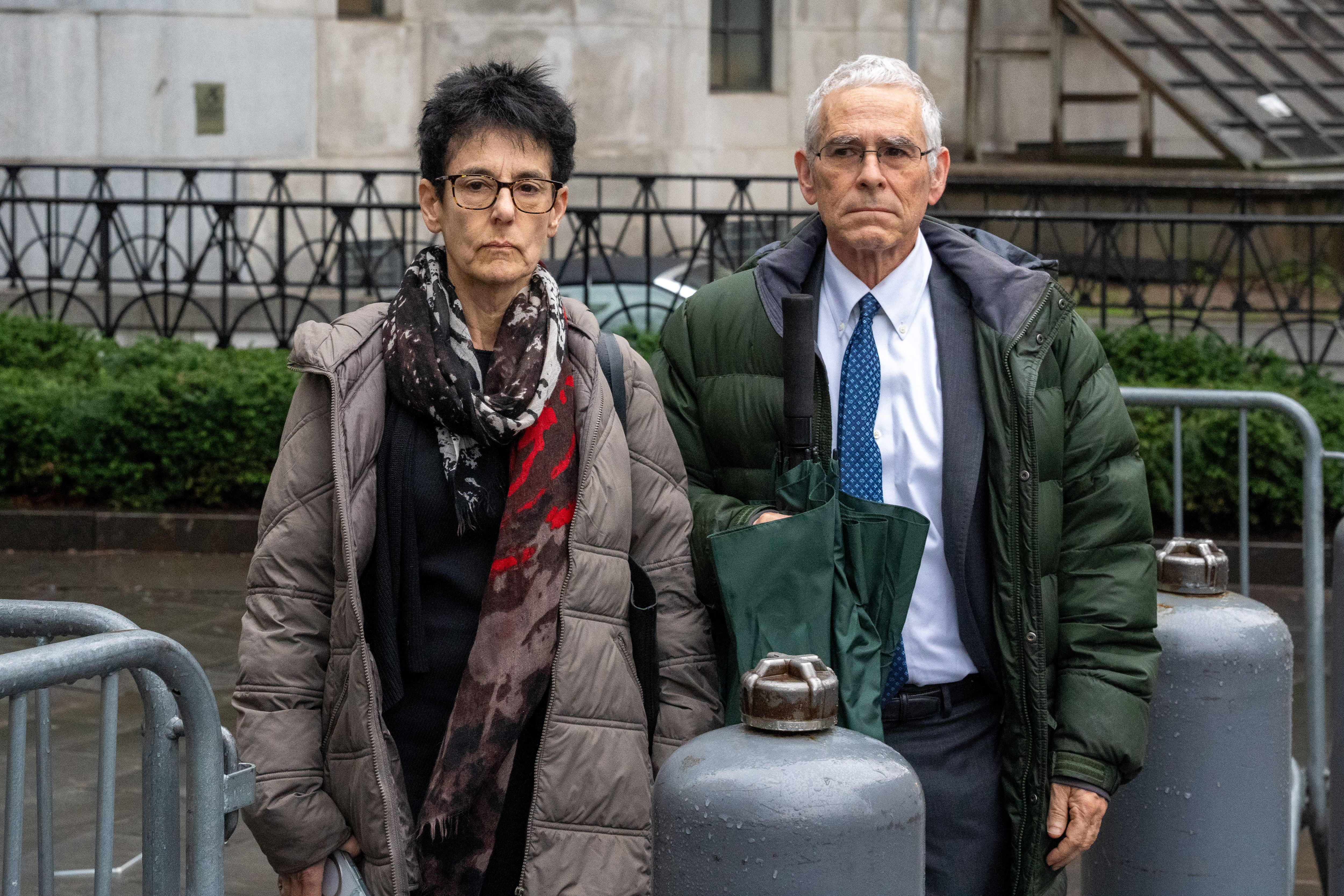 A gray-haired elderly man and a elderly woman with black hair, both wearing glasses, stand outside on a city street on a rainy day while wearing puffy coats.