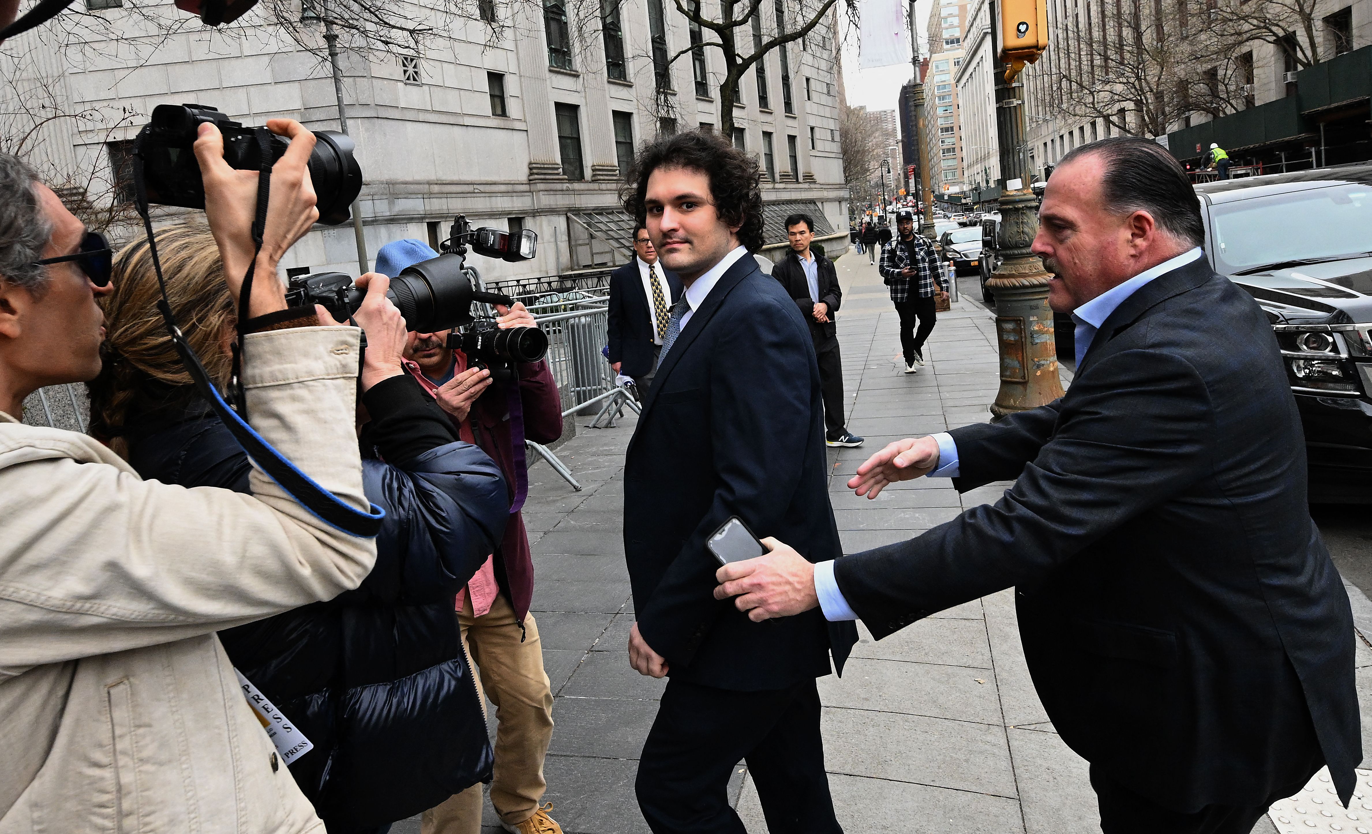 A clean-shaven man with dark curly hair, wearing a dark suit and tie, looks into the camera as photographers and reporters surround him on a New York City sidewalk. A man in a suit behind him holds up his hands, as if attempting to clear a path.