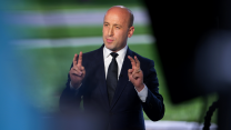 A bald man in a suit makes air quotes with his fingers while speaking.