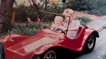 A photo of a toddler and her older brother seated next to one another in a red car on a track that’s laid through a thicket of flowers and bushes. The car, designed for children, is based on an early 20th century racing car.