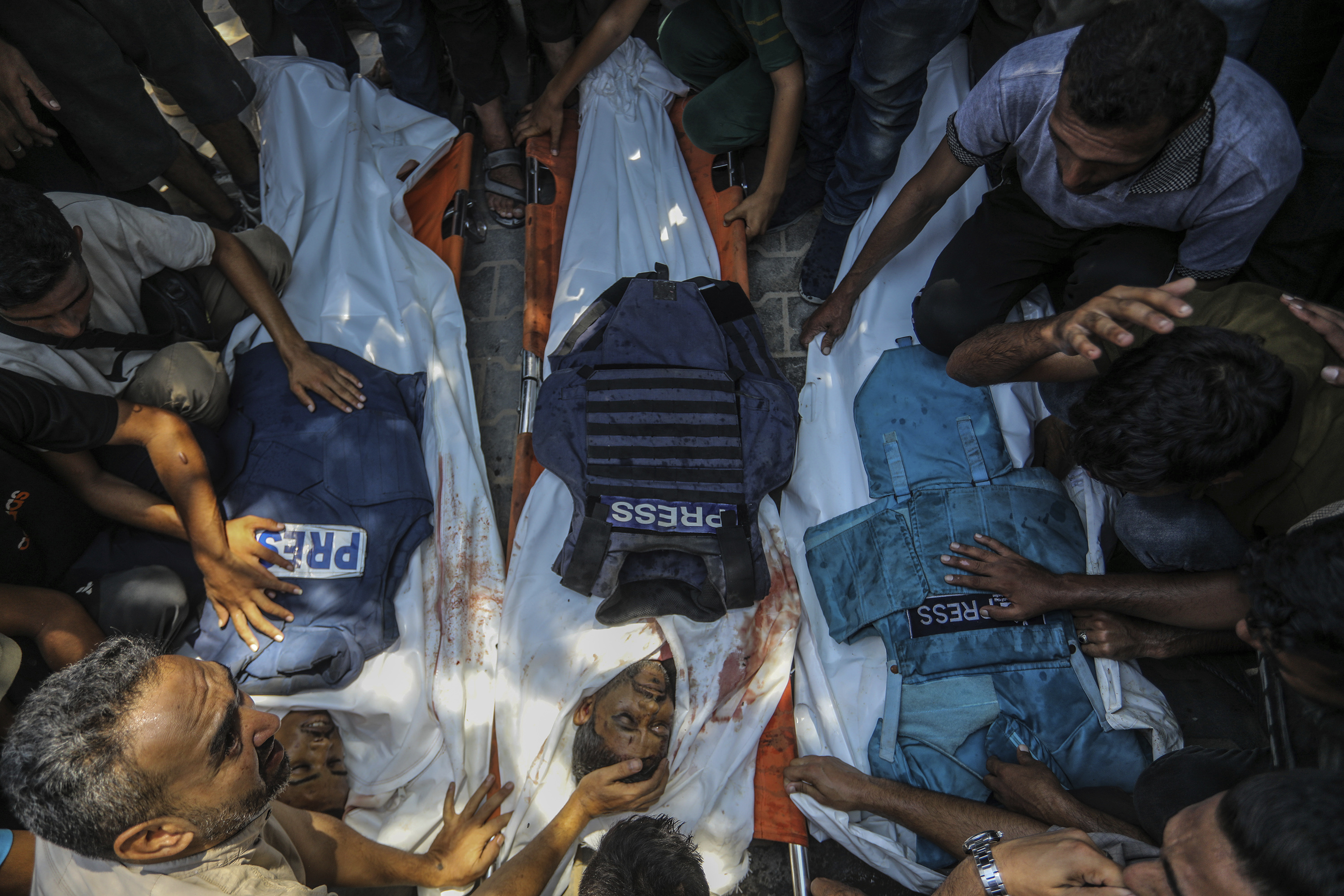Overhead shot of a group of mourners grieving the shrouded bodies of three Palestinian journalists—their press vests lay atop the shrouds.