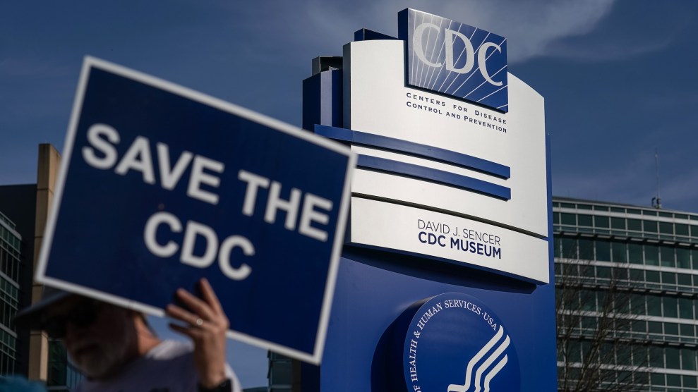 A person holds a "Save the CDC" poster in front of a large blue and white CDC sign on CDC campus