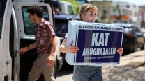 a woman holding a sign in front of a Kat for Congress sign on a street