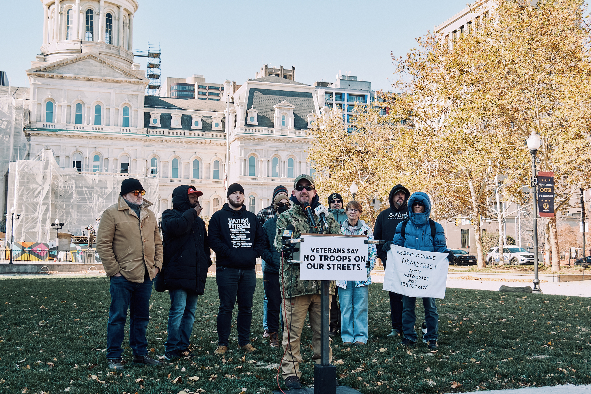 Buzz Grambo, wearing a baseball hat, camouflage jacket, tan pants, and sunglasses, on a podium in a small park. The podium has a sign that reads, "Veterans say no troops on our streets." Behind him is a small group of people, with one person holding a sign that reads, "We served to ensure democracy, not autocracy, not plutocracy."