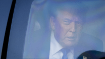 A close-up photograph of Donald Trump peering out a back window of the presidential limousine. With narrowed eyes and mouth slightly ajar, he is wearing a suit and tie with an American flag pin on his left lapel.