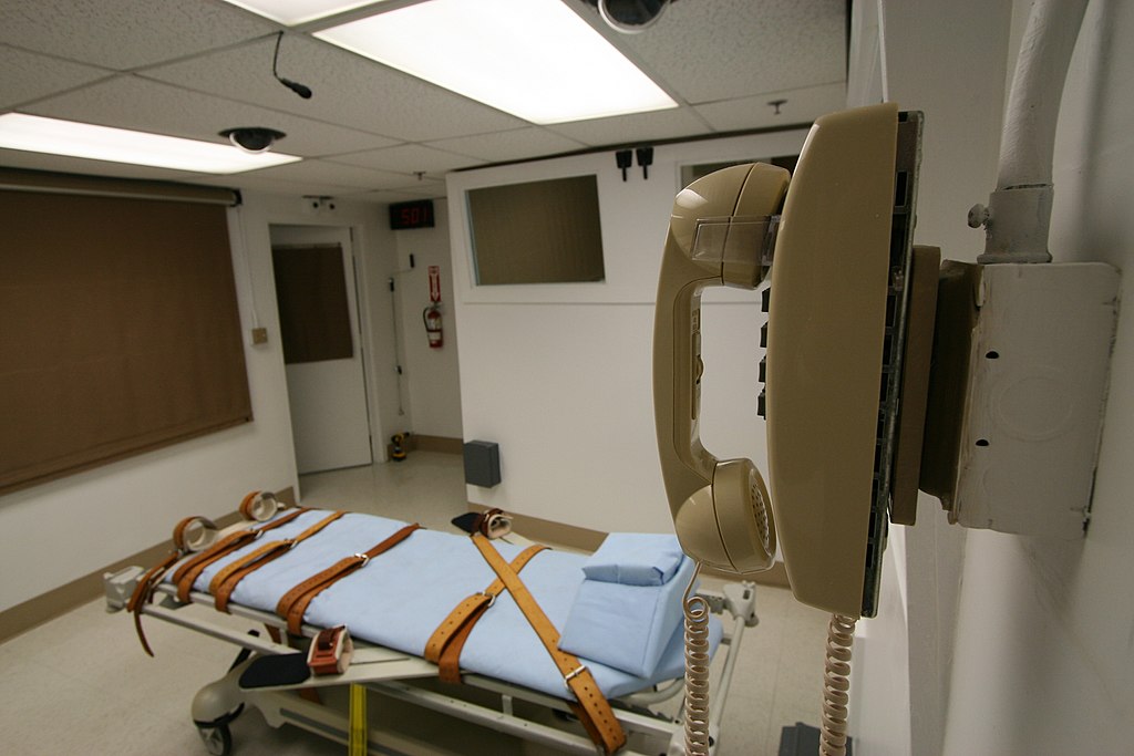 A photo of a sterile room that is empty except for a table with brown straps and a tan phone attached to the wall.