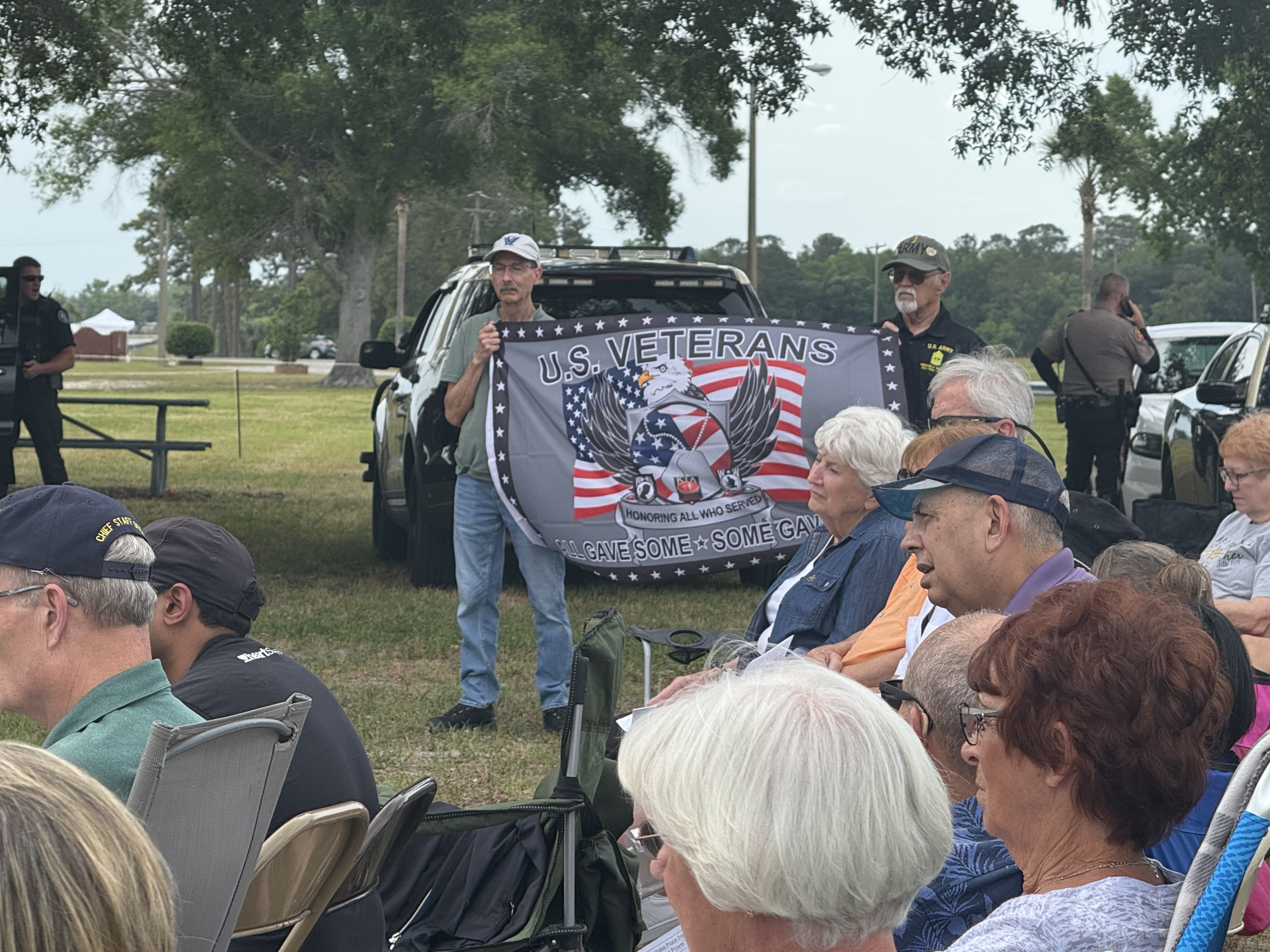 A photo of a crowd of mostly elderly people siting in lawn chairs. Two men, one with a mustache, another with a white goatee, hold a flag that reads "U.S. Veterans: All Gave Some. Some Gave All."