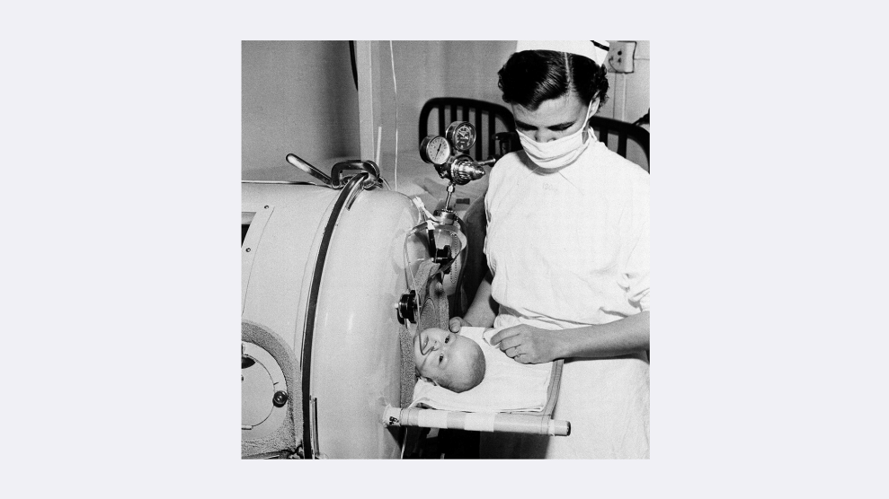 A photo of an infant's head lying on a prop, as the rest of her body is contained within an iron lung. A female nurse dressed in white standing over the child looks at her attentively.