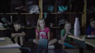 A darkly lit photo of an elementary school classroom with children staring into the distance pensively, as they sit on the floor with their backs against a wall of backpack compartments.