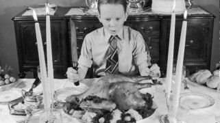 circa 1955, a black and white image of a redheaded boy, maybe 11 or so, sitting in shirt and tie at a table with a roast turkey in front of him, holding carving implements and looking very excited