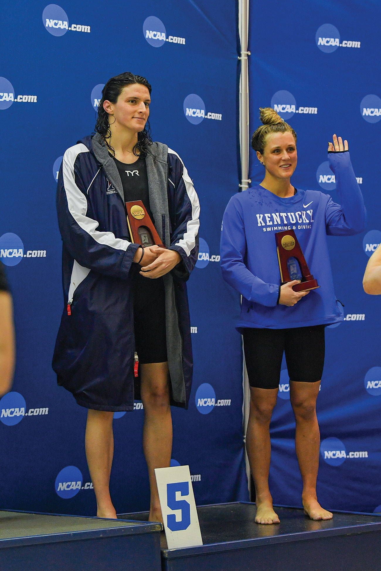 Two athletes in swim team uniforms stand on a podium holding NCAA trophies, positioned in front of a floor marker displaying the number 5.