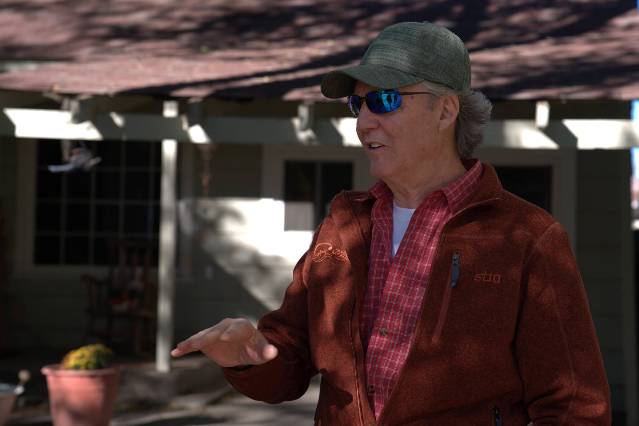 A man wearing a baseball cap, sunglasses, and a red jackets speaks in front of a house.