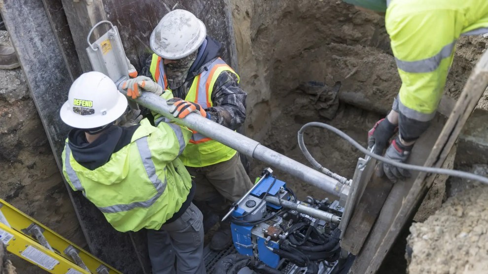 Workers wearing hard hats and yellow protective vests work on a pipe.