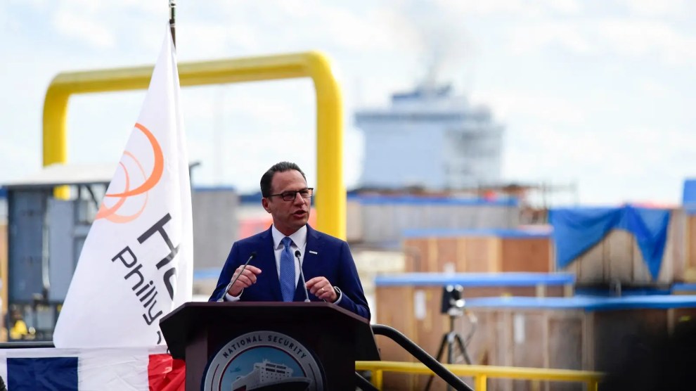A white man with a blue suit stands at a podium in front of a ship yard.