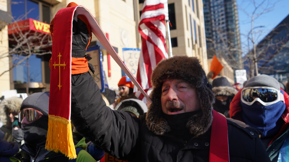 An elderly man with a dark mustache dressed in a black winter coat and fur-lined hat holds up a red stole with a gold cross. He stands amid other protesters on a city street under a blue sky.