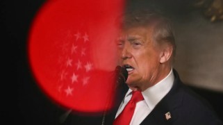 Trump, an elderly man wearing a red tie, stands in front of a red circle.