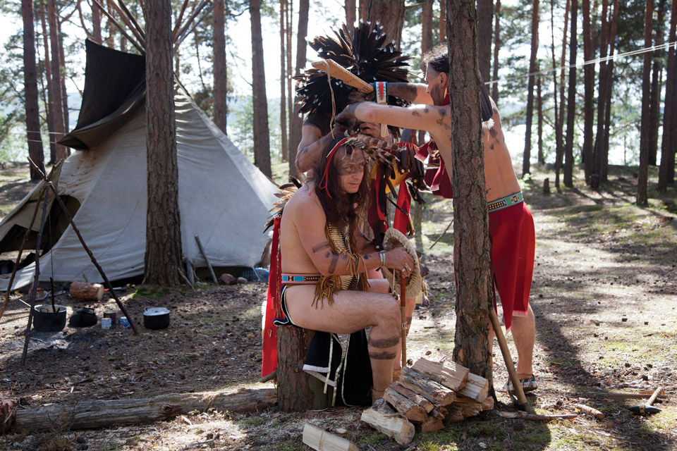 Man dressed as Native American being fitted with feather headdress.